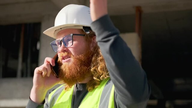 Indignant builder argues on mobile phone, gesturing with hand, discussing mistakes in planning construction work, standing against the backdrop of an unfinished building in a hard hat on a sunny day.
