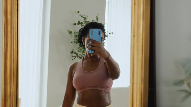 Young black woman doing a workout at home, takes a break to rest and uses a mobile phone. Uses online app for training and chatting. Smiles and feels contented and pleased, taking selphie photo