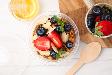 Tasty granola with berries, almond flakes and mint in plastic cup on white wooden table, flat lay