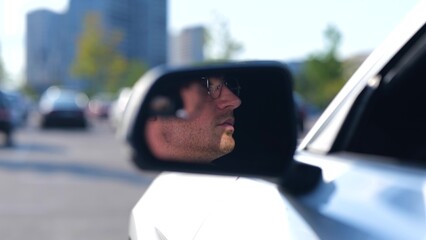 Happy young man sitting in the car while looking on the side view mirror, wearing glasses. Transport and lifestyle concept. Real time