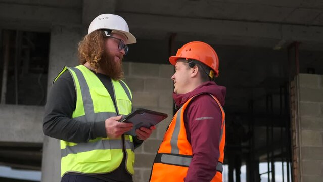 Civil architect engineer bearded man and builder worker in orange helmet discussing work blueprints in digital tablet and working outdoors on structure construction site.