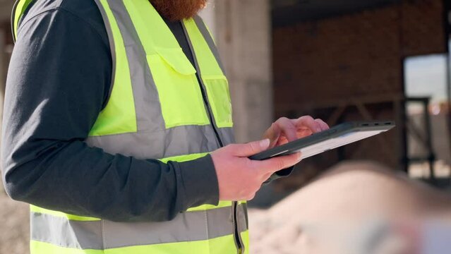 Close-up of hands holding digital tablet of professional architect engineer expert assessing progress of building's construction while typing with his finger on the screen of device. 