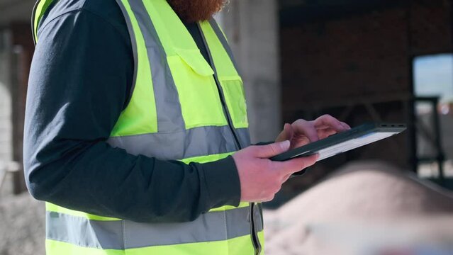 Unknown worker man hands hold tablet computer at construction site in safety vest, close up using modern gadget pad. Close up.
