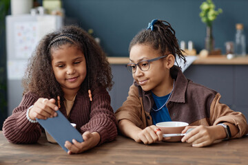 Front view portrait of two African American teenage girls looking at smartphone screen while sitting at table in kitchen - Powered by Adobe