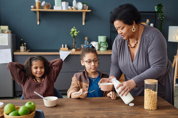 Portrait of caring African American grandma serving breakfast to two young girls at kitchen table