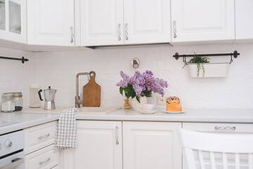 Spring morning in light kitchen: bouquet of lilac flowers, cake, kulich, glass of coffee bowl of sugar in white kitchen.
