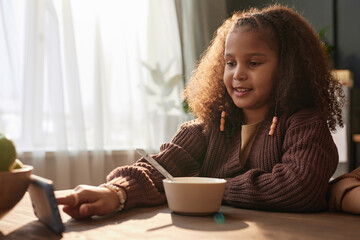 Portrait of teen African American girl using phone at kitchen table during breakfast at home scene...