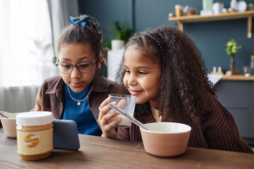 Portrait of two African American girls watching videos via smartphone at kitchen table during...