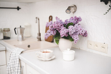 Spring morning in light kitchen: bouquet of lilac flowers, glass of tea, bowl of sugar in white kitchen.