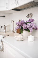 Spring morning in light kitchen: bouquet of lilac flowers, glass of coffee, bowl of sugar in white kitchen.