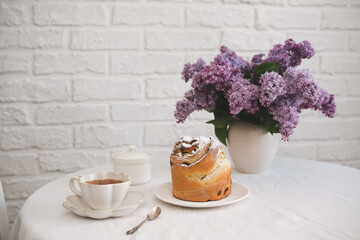 Spring morning in light living room: bouquet of lilac flowers, cake, kulich, glass of green tea, bowl of sugar in white bricks background.
