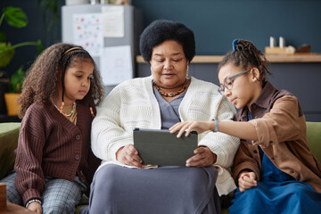 Portrait of two young girls teaching grandmother using tablet at home sitting on couch together