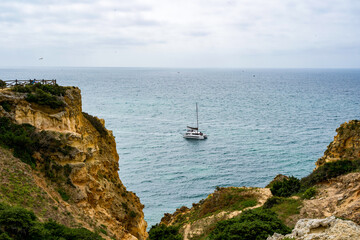 White sailboat in Atlantic ocean of Portugal and seacoast with yellow rocks and green grass....