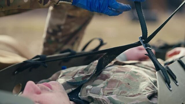 Close up a confident male army medic in a camouflage uniform and rubber blue gloves quickly unties the straps on a green stretcher for wounded soldiers during training of Medics and soldiers on the