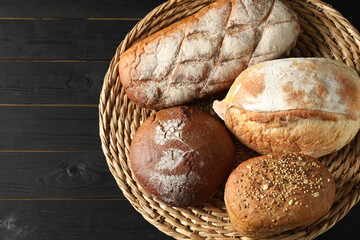 Wicker basket with different types of fresh bread on black wooden table, top view