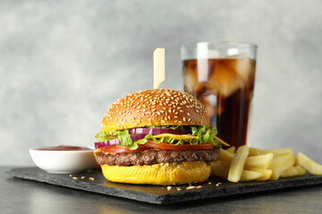 Burger with delicious patty, soda drink, french fries and sauce on dark table against gray background, closeup