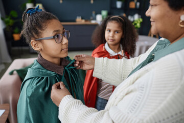 Side view of Black senior woman helping smiling young girl wearing costume cape at home