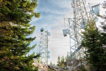 A group of broadcast antennas on a mountain top among fir trees against the sky.