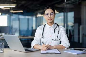 A focused and thoughtful Asian female doctor with glasses sits at her office desk, equipped with a laptop and medical implements, contemplating her next steps in patient care.