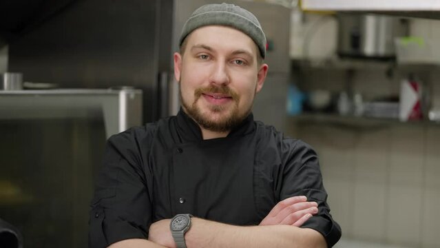 Portrait of a happy chef with a beard in a black uniform who folds his arms on his chest and poses for the camera in the kitchen in a restaurant