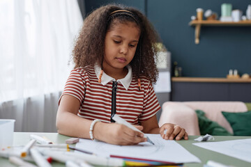 Portrait of little Black girl drawing picture with pencils sitting at table at home, copy space