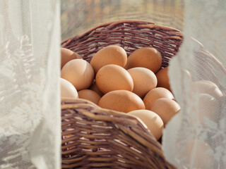 Vertical close up shot of a basket full of eggs on a windowsill behind white laced curtains. Each...