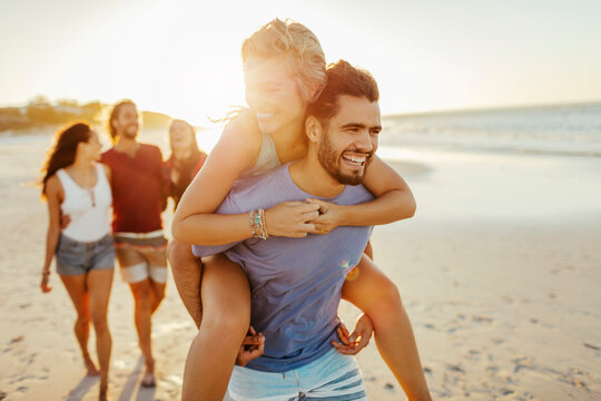 Young couple enjoying a piggyback ride on the beach with friends in the background