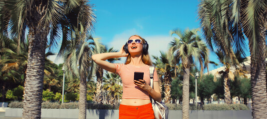 Happy smiling young woman listening to music in headphones with phone in summer park with palm trees