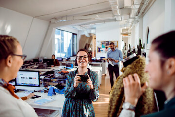 Smiling female fashion designer holding a camera in a lively design studio with colleagues...