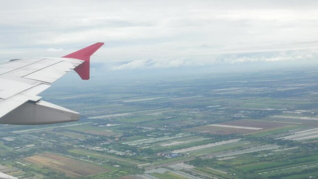 view through aircraft window with plane's wing over green farm field site suburb of Bangkok area and airplane aircraft make a turn in sky