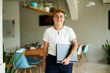 Confident transgender person with tattoos holds laptop, smiles in modern eco office. Inclusive...