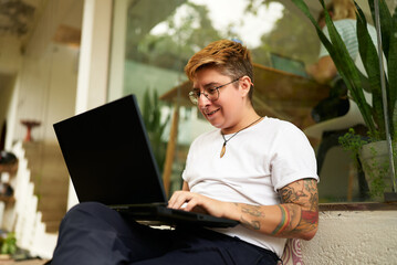 Tattooed Gen Z transgender person works on laptop in cozy office space, displays casual, inclusive...