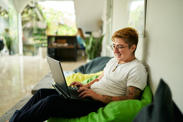 Transgender Gen Z individual smiles while working on laptop seated on green bean bag in airy...