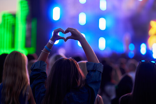 Person forms heart shape with hands at lively outdoor music festival, blurred stage lights, crowd enjoys event. Love symbol gesture, fans celebrate artist, youth culture, happy summer vibe in concert.