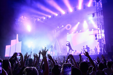 Silhouettes of excited crowd at music fest, hands raised with lit stage in background. Band...