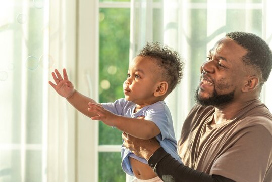 Happy african black parents dad father throw baby son on piggyback and neck riding teasing fun near window. Black baby son and daddy enjoy teasing kiss cheek and neck riding at window light
