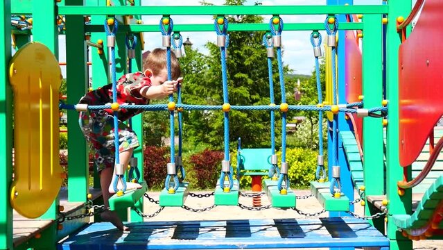 A little cute boy climbs onto the logs of a playground bridge hanging on chains and walks along them