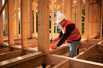 Carpenter constructing wooden frame house. Bearded man with glasses is hammering nails into the structure, wearing protective helmet and construction vest. Concept of modern ecological construction.