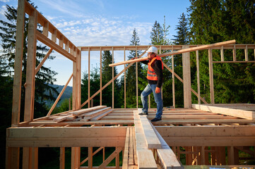Carpenter building wooden-framed house near the forest. Man holds large board, dressed in work clothes and helmet. Concept of contemporary and eco-friendly construction.