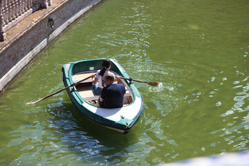 Obraz premium two tourists, a man and a woman, enjoy the sunny day rowing in the boats on the canal in the square of spain in seville. Travel concept and holiday destination.