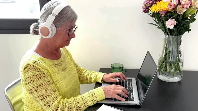 Concentrated retired senior woman wearing headphones while typing on laptop, beautiful white haired lady loves technology and social media, creates blog postcast