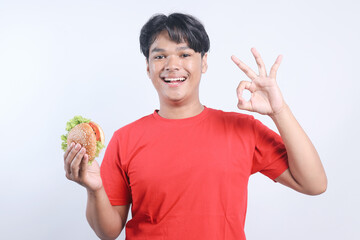 Happy Young Asian Man While Holding Burger And Showing Ok Sign Isolated On White Background 