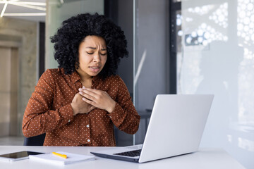 African American woman in a polka dot shirt experiences discomfort and chest pain while working on...