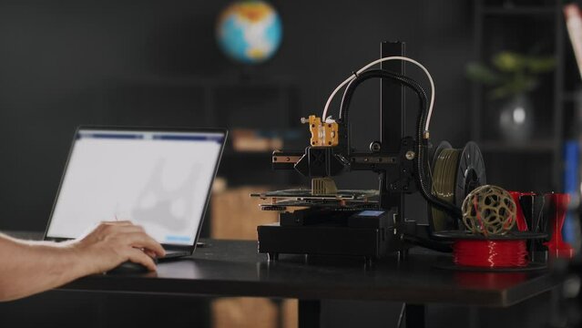 A graphic designer uses a laptop and makes a sketch of postoperative fixation bandage in red colour for the heart and chest for women, depicted on a device screen. A 3d printer machine works in