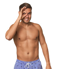 Young man shirtless in swimwear, studio excited keeping ok gesture on eye.
