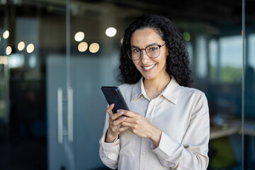 Cheerful professional woman with curly hair, wearing glasses, smiles while using a smartphone in a modern office setting. The image conveys positivity and communication.