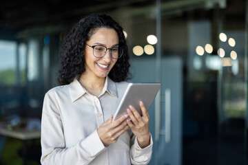 A cheerful professional woman with curly hair smiles as she uses a tablet in a bright, modern office setting. The image conveys engagement and productivity.