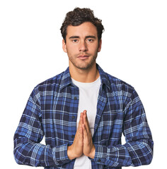 Young Hispanic man in studio praying, showing devotion, religious person looking for divine...