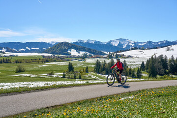active senior woman riding her electric mountain bike on a sunny day in  spring with Dandelion flowers on the meadows below snow capped mountains of Nagelfluh chain near Oberstaufen, Allgaeu, Germany