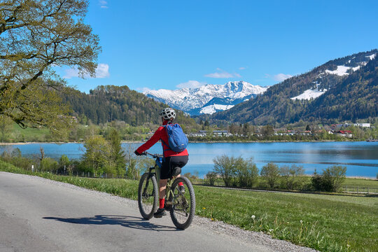 nice senior woman riding her electric mountain bike in springtime at lake Alpsee near Immenstadt, with snowcapped High Algaeu mountains in Background,Allgau,Bavaria, Germany